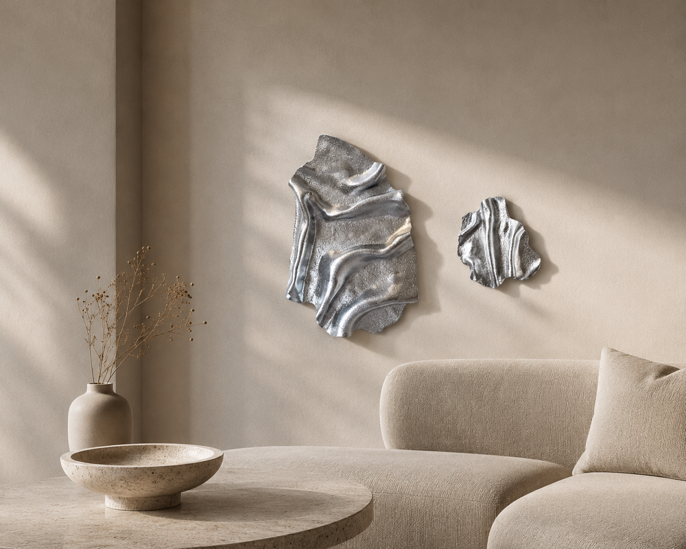 To håndlavede, skulpturelle vægværker i sølv ophængt side om side på en lys, minimalistisk væg over en beige sofa. Det store værk måler 40 × 28 cm, og det mindre måler 22 × 25 cm. De organiske former og den metalliske overflade skaber et eksklusivt udtryk i et moderne, skandinavisk interiør.