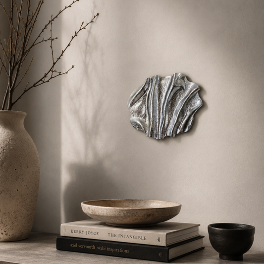Unikt håndlavet vægværk i sølv med skulpturel tekstur og metallisk finish placeret i et roligt, minimalistisk miljø med bløde skygger og elegante boligdetaljer.