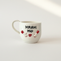 Håndlavet hvidt porcelænskrus med “KRAM MIG”-tekst, små hænder og rødt hjerte fotograferet i lyst studiemiljø.